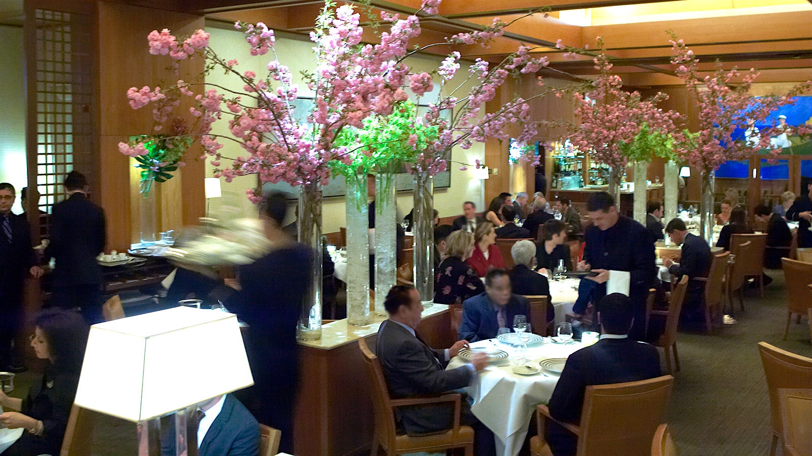People dining in Le Bernardin’s dining room with a coffered ceiling and slender vases of flowers throughout